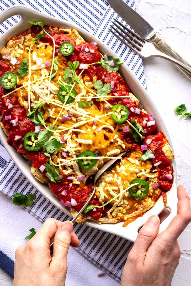 Overhead shot of Black Bean Vegan Enchiladas. A woman's hand is spooning an enchilada out of a casserole dish. It's topped with jalapeno slices and red onion, and is on a blue and white striped tea towel.  