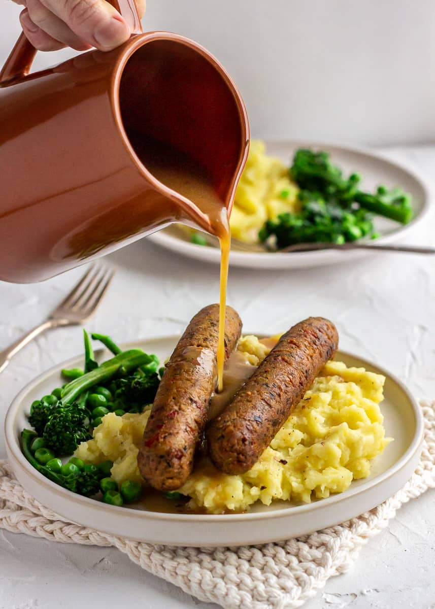 An orange jug is seen pouring vegan gravy over a plate of vegetarian sausages, mashed potatoes and vegetables. There is another plate of food in the background and the whole scene is very bright.
