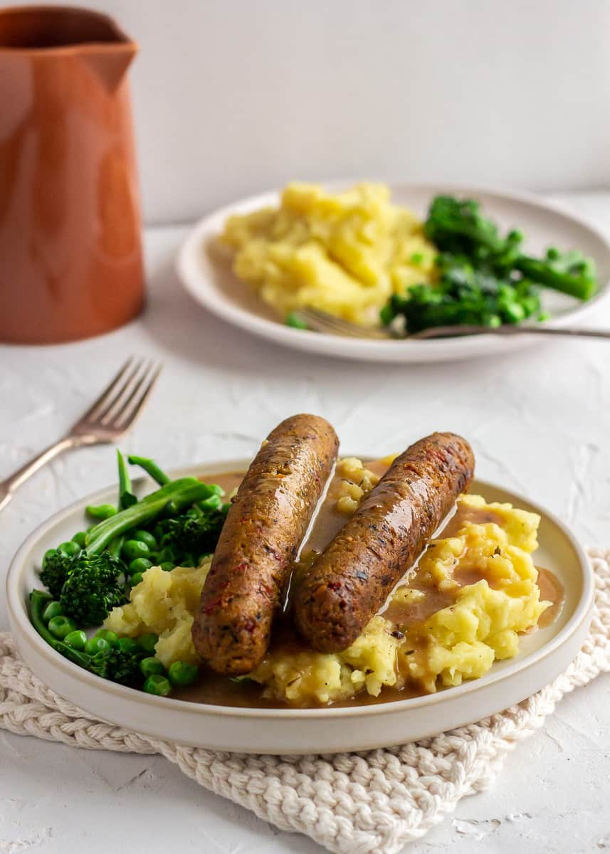An orange jug is seen next to two plates of vegetarian sausages, mashed potatoes, vegetables and vegan gravy. The whole scene is very bright and the plates are on a white textured background.