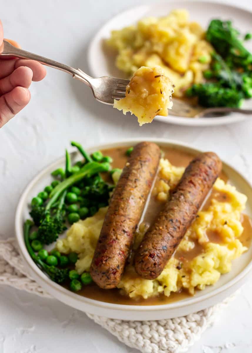 A hand lifts a forkful of mashed potatoes covered in gravy over a plate of veggie sausages and vegetables.