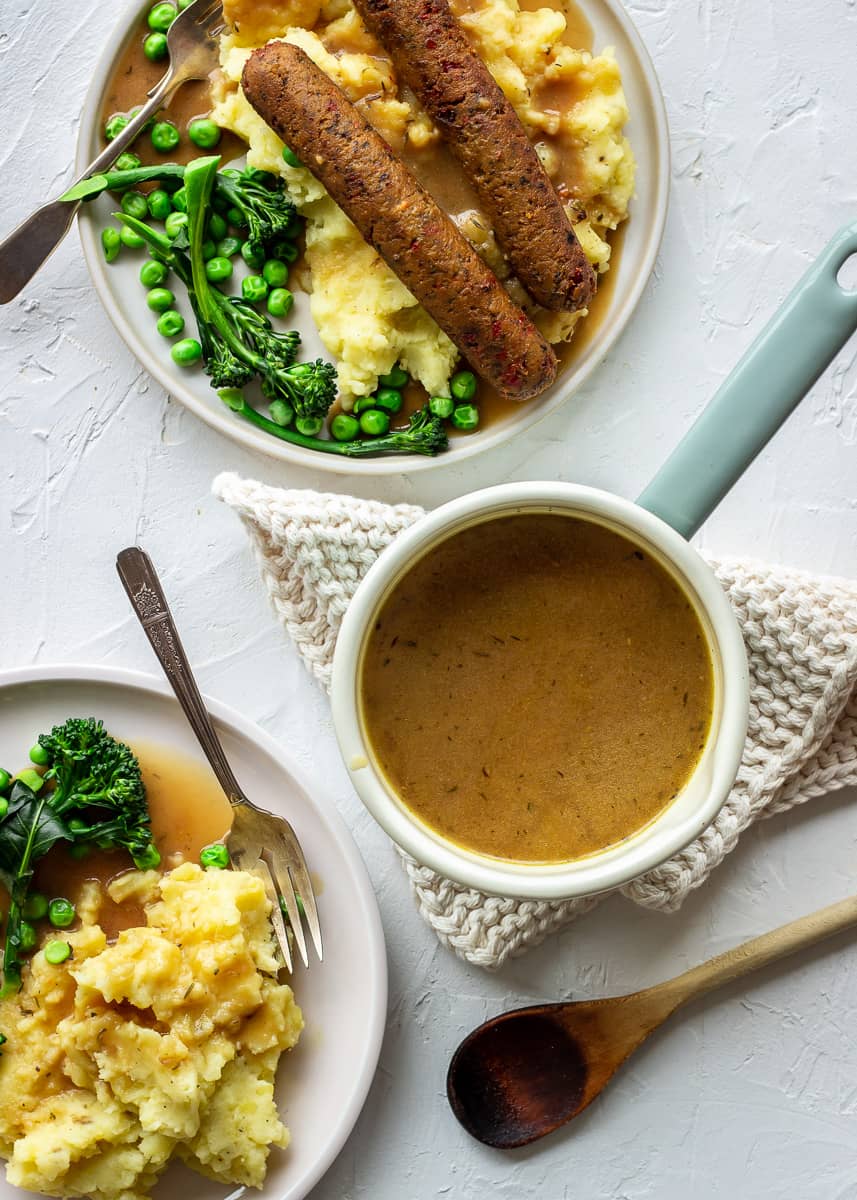 Overhead shot shows a saucepan of vegan gravy surrounded by two plates of vegetarian sausages, mashed potatoes and vegetables.