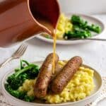 A woman's hand pours a jug of vegan gravy on to plate of sausages and mashed potato.