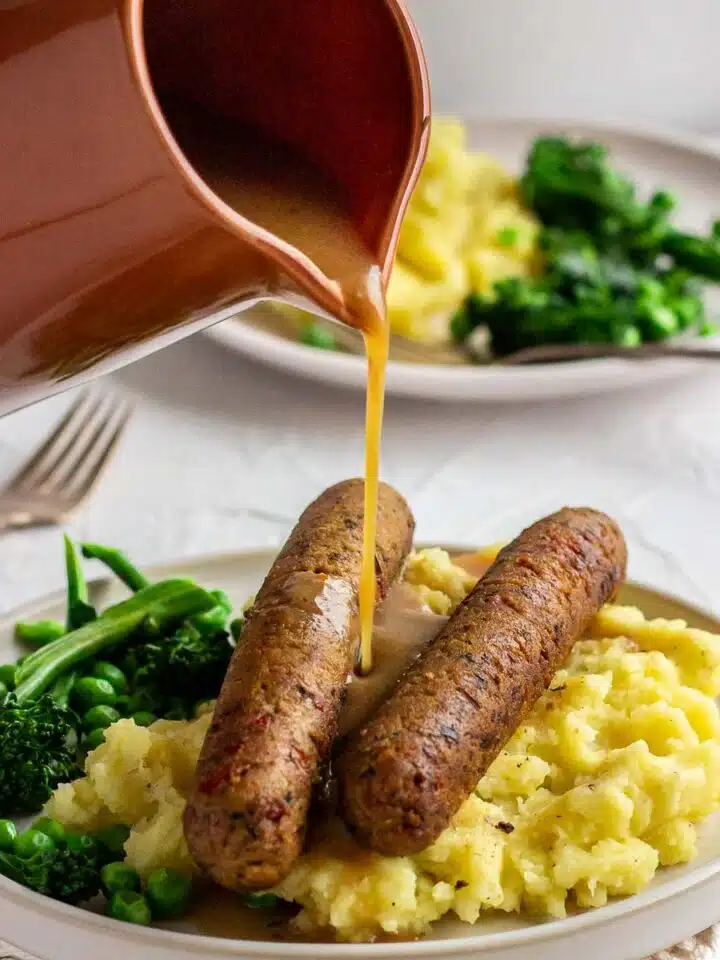A woman's hand pours a jug of vegan gravy on to plate of sausages and mashed potato.