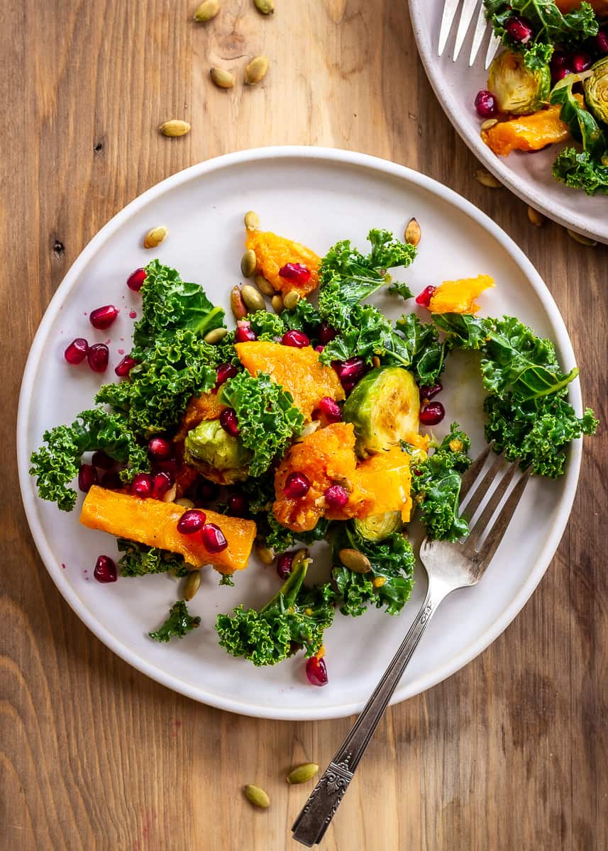 Overhead image of Brussels sprout salad with roasted butternut squash, kale, pumpkin seeds and pomegranate. Salad is on a grey plate with a silver fork. The plate sits on a neutral wooden background.
