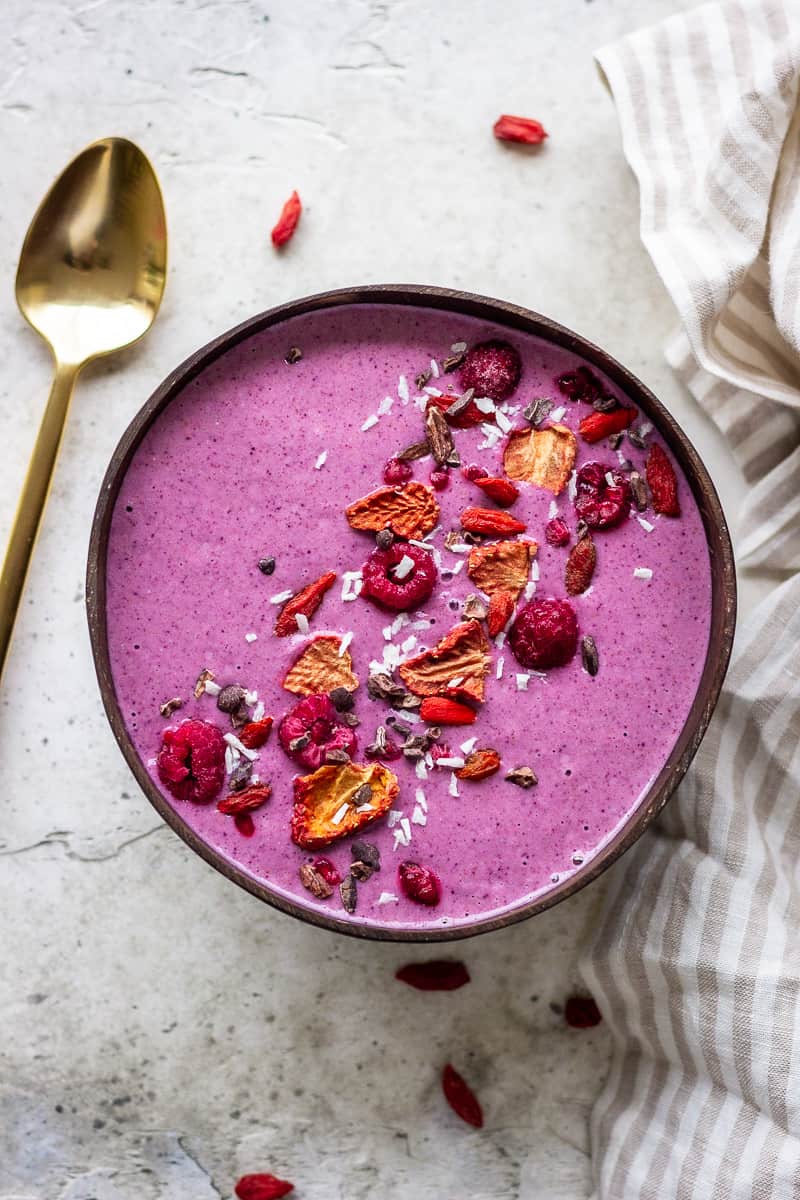Purple berry smoothie bowl decorated with raspberries, freeze-dried strawberries, coconut and cacao nibs. Next to the bowl is a golden spoon and a neutral striped napkin.