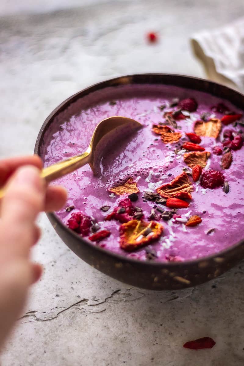A woman's hand stirs a purple berry smoothie bowl with a gold spoon. The smoothie is decorated with strawberries, raspberries and cacao nibs.