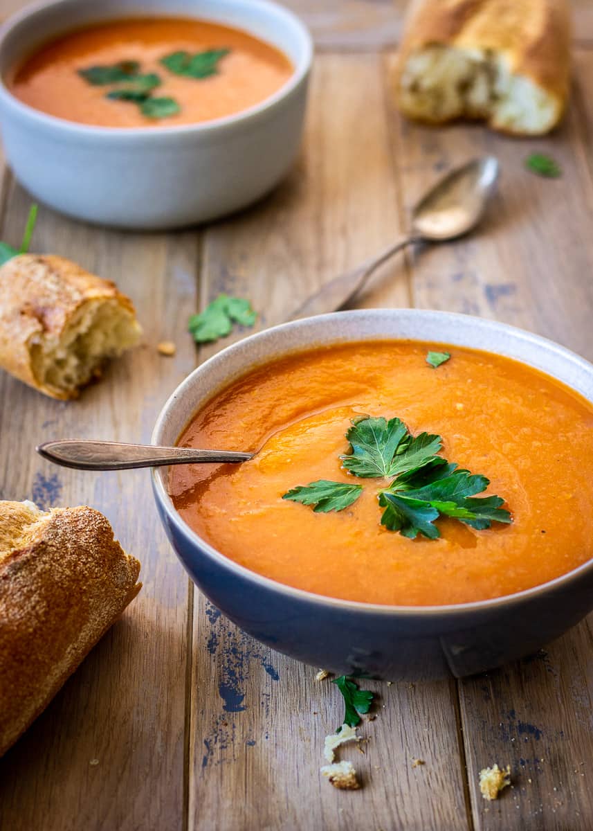 A bowl of Carrot, Ginger & Apple Soup sits on a wooden background. It is surrounded by bread and is decorated with parsley leaves. There is another bowl of soup in the background.
