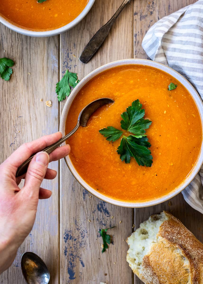 A bowl of Carrot, Apple & Ginger Soup with a woman's hand taking a spoonful from the bowl.