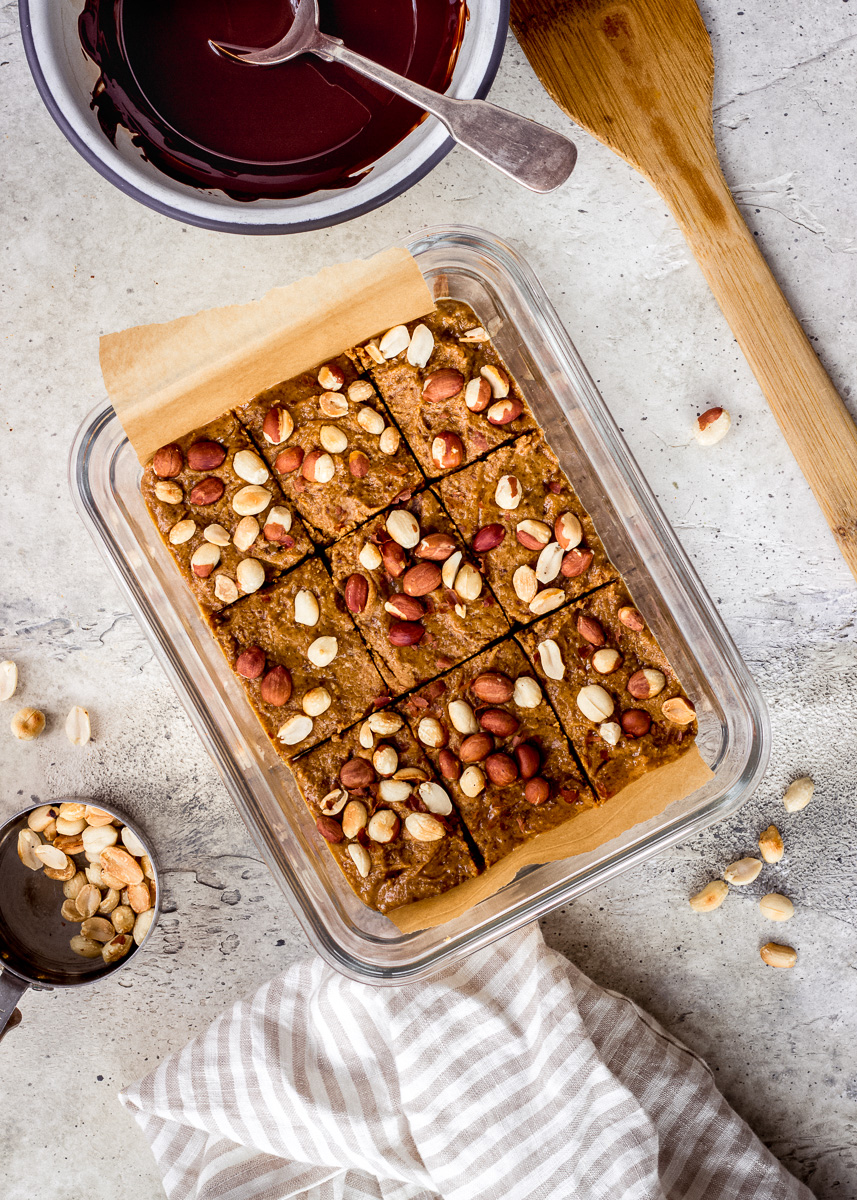 Overhead view of peanut butter bars in a glass food storage container, decorated with roasted peanuts.