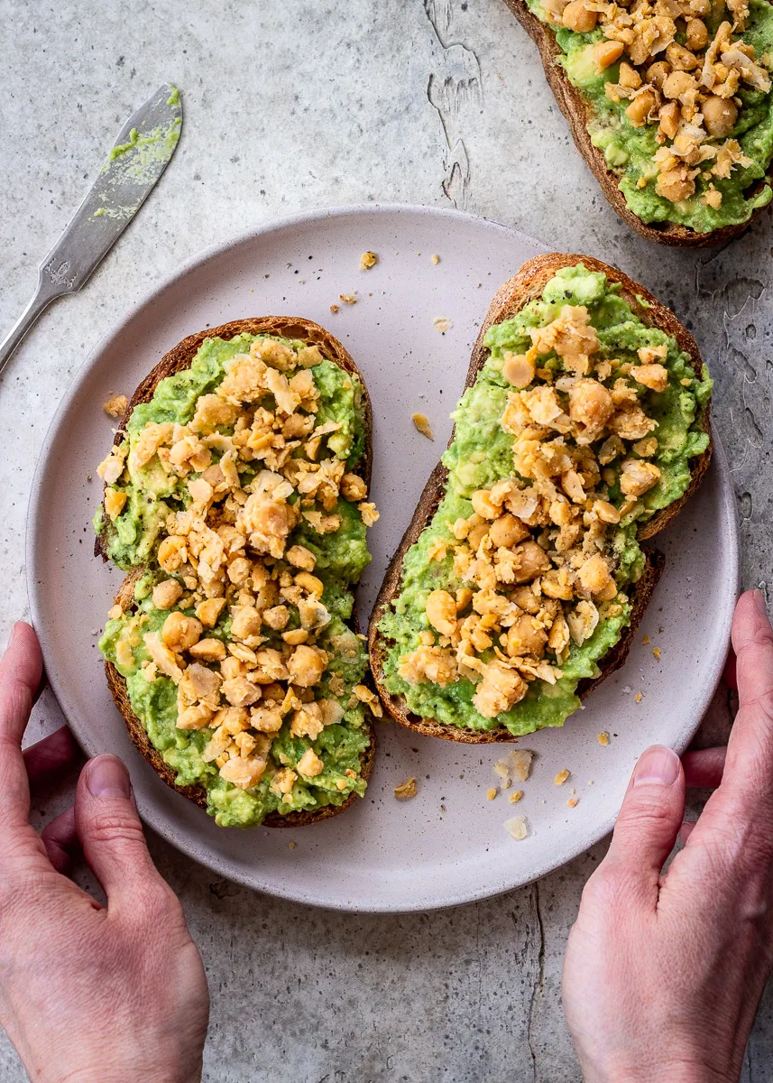 A woman's hands holding a plate of toast with avocado and smashed chickpeas.
