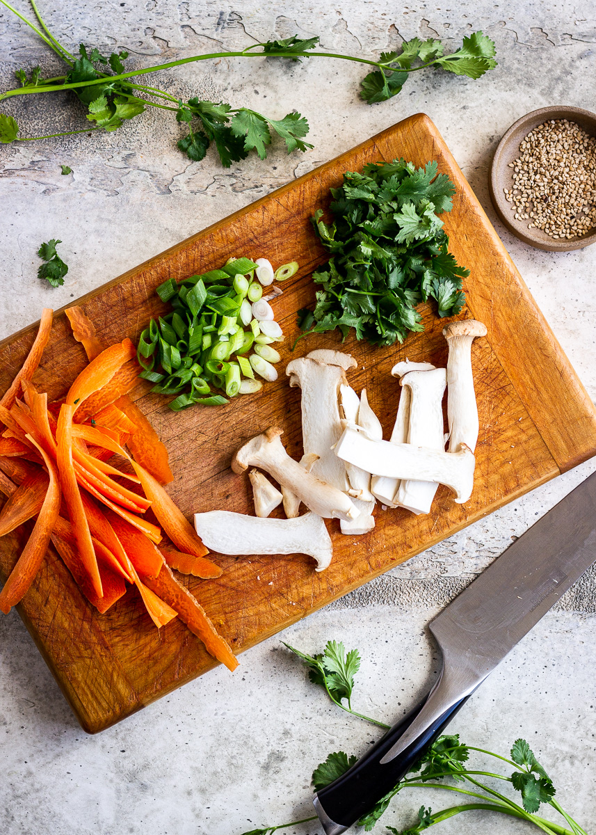 Sliced mushrooms, carrots, green onions and cilantro on a chopping board.