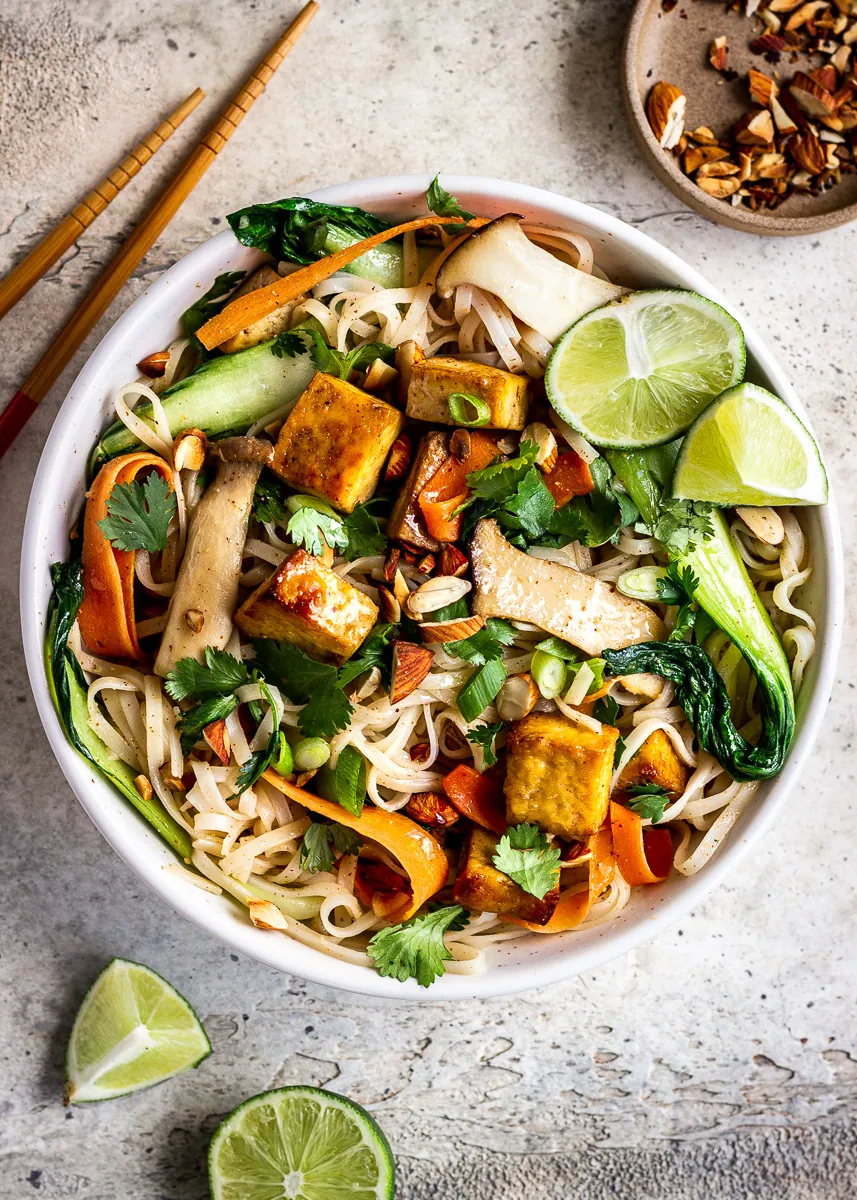 A bowl of noodles, tofu and vegetables with almonds, chopsticks and lime wedges nearby.