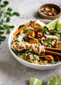 A bowl of noodles, tofu and vegetables with almonds, cilantro and lime wedges nearby.