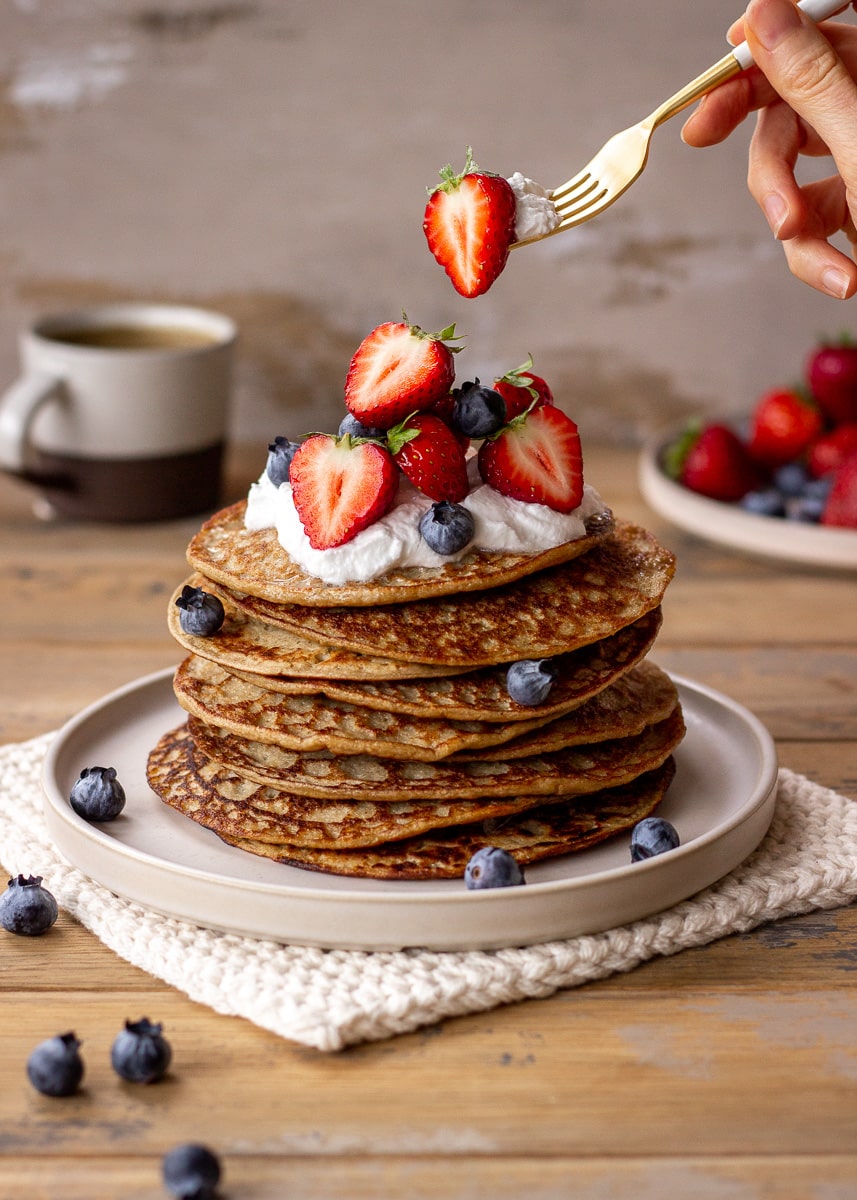 A stack of 3 ingredient banana oatmeal pancakes topped with coconut whipped cream and berries, with a woman's hand holding a fork.