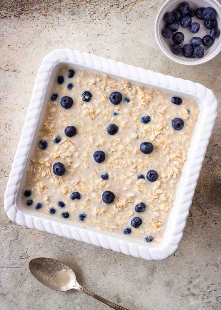 Vegan baked oatmeal in white dish scattered with blueberries, ready to be cooked.