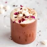 Glass mug of rose hot chocolate topped with whipped cream, rose petals and marshmallows.