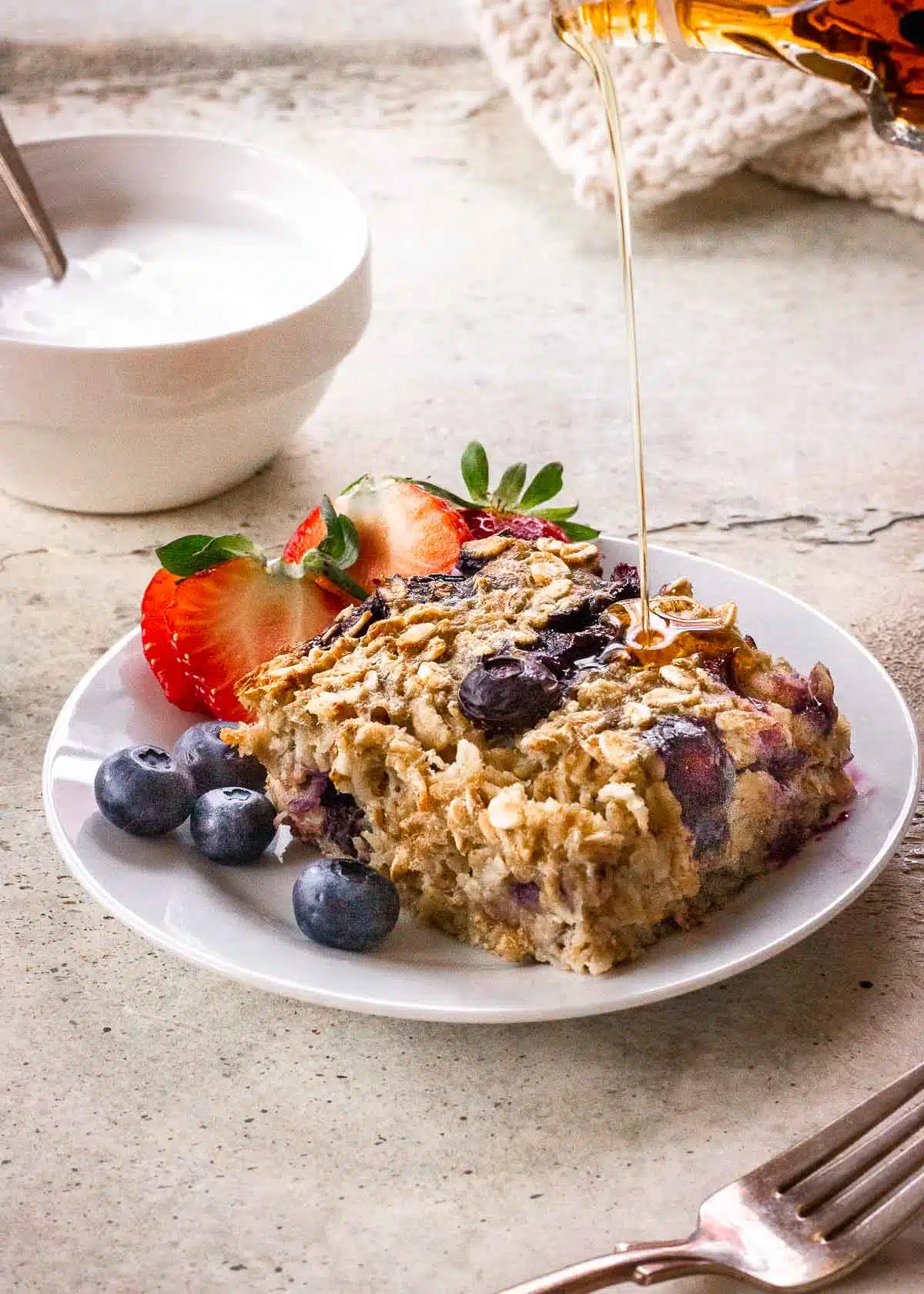 A slice of vegan baked oatmeal on a plate surrounded by strawberries and blueberries, and being drizzled with maple syrup.