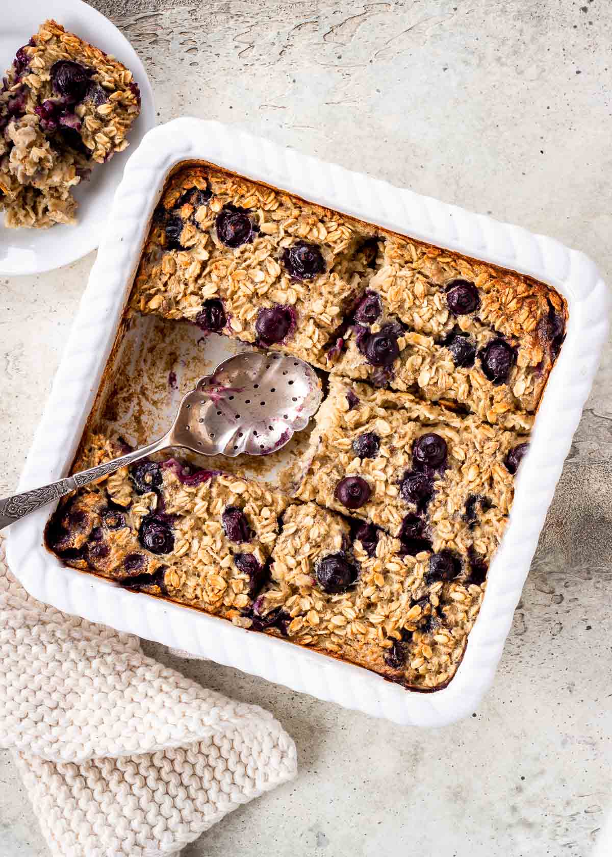 A large white tray of vegan baked oatmeal cut into slices and studded with blueberries. A spoon has already taken a slice out.