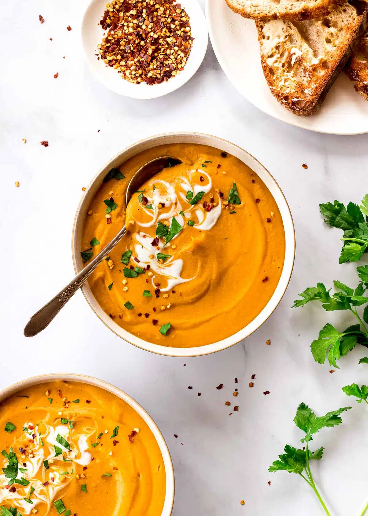 Two bowls of Carrot and Lentil Soup surrounded by chili flakes and fresh bread.