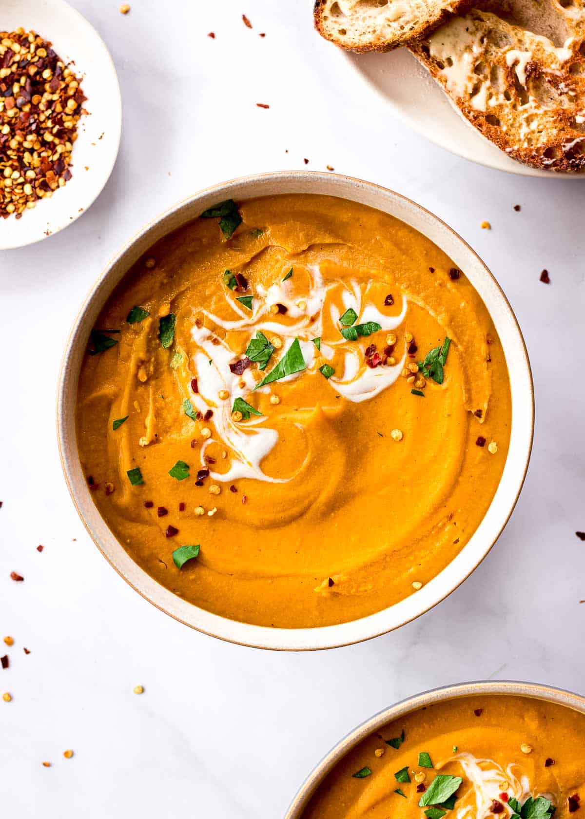 A bowl of lentil and carrot soup deorated with coconut yoghurt, parsley and chili flakes surrounded by bread and chili flakes.