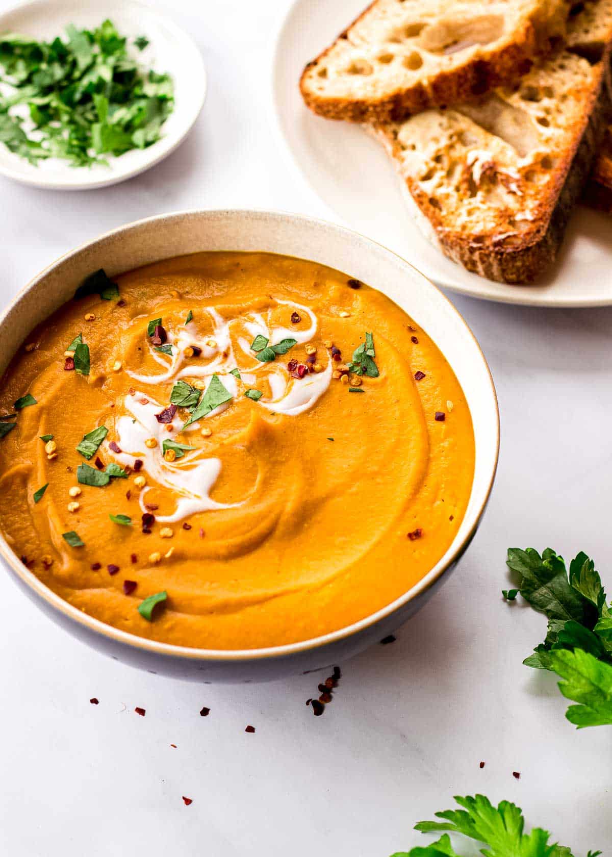 A bowl of lentil and carrot soup deorated with coconut yoghurt, parsley and chili flakes surrounded by bread and parsley.