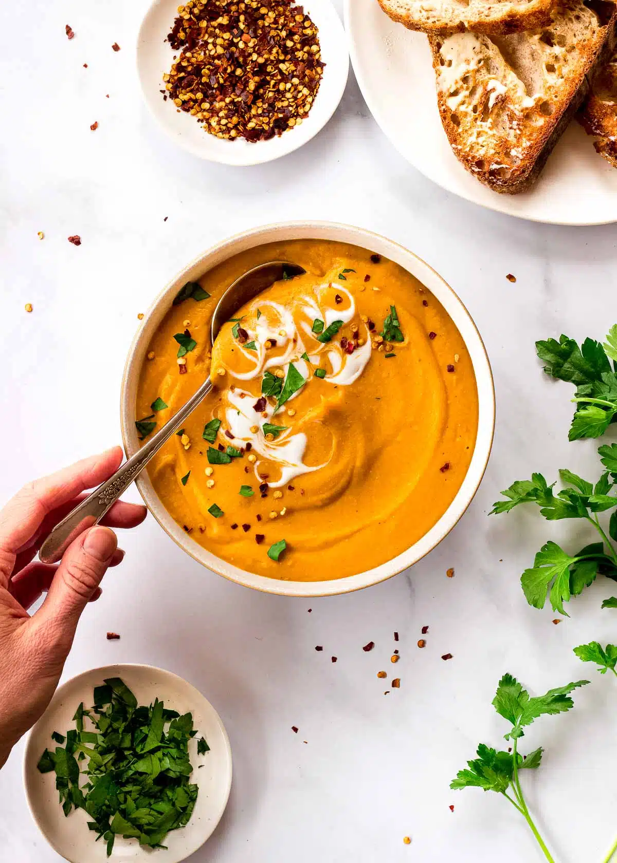 A woman's hand reaches for a spoon in a bowl of Lentil and Carrot Soup decorated with yoghurt and parsley.