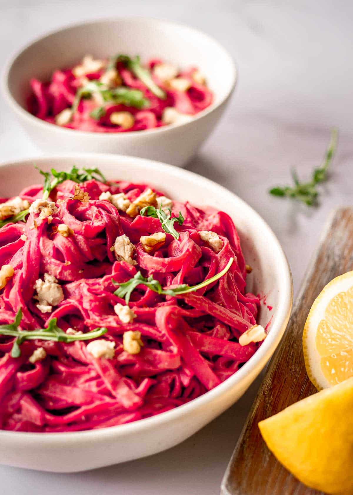 Two white bowls of beet pasta decorated with arugula and walnuts.