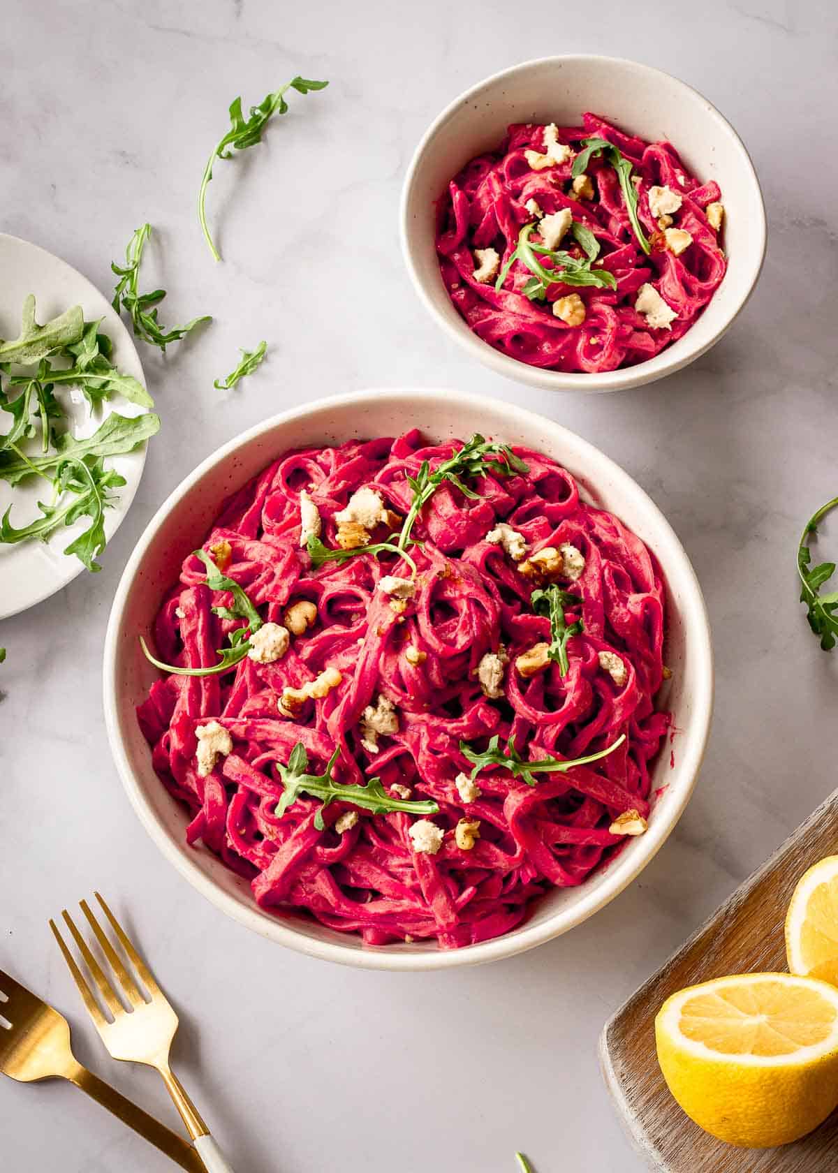 Two white bowls of pasta with beet sauce decorated with arugula, vegan cheese and walnuts.