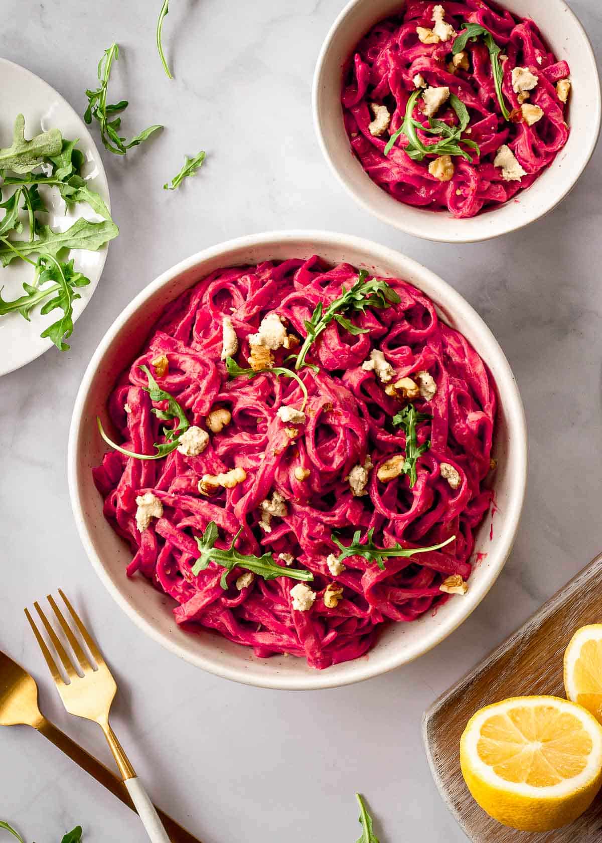 Two white bowls of beet pasta decorated with arugula and walnuts, with lemon halves on the side.