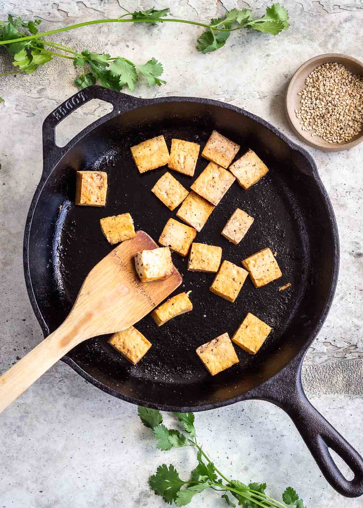 Tofu in a cast iron pan being flipped with a wooden spatula.