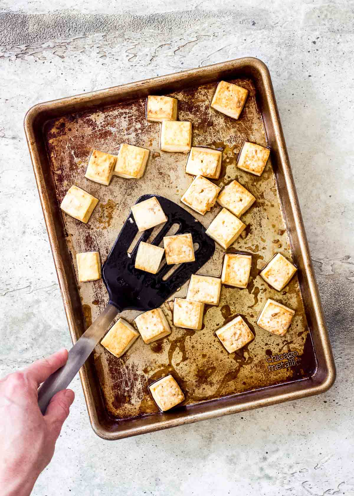 Tray of crispy broiled tofu being flipped with spatula by female hand.