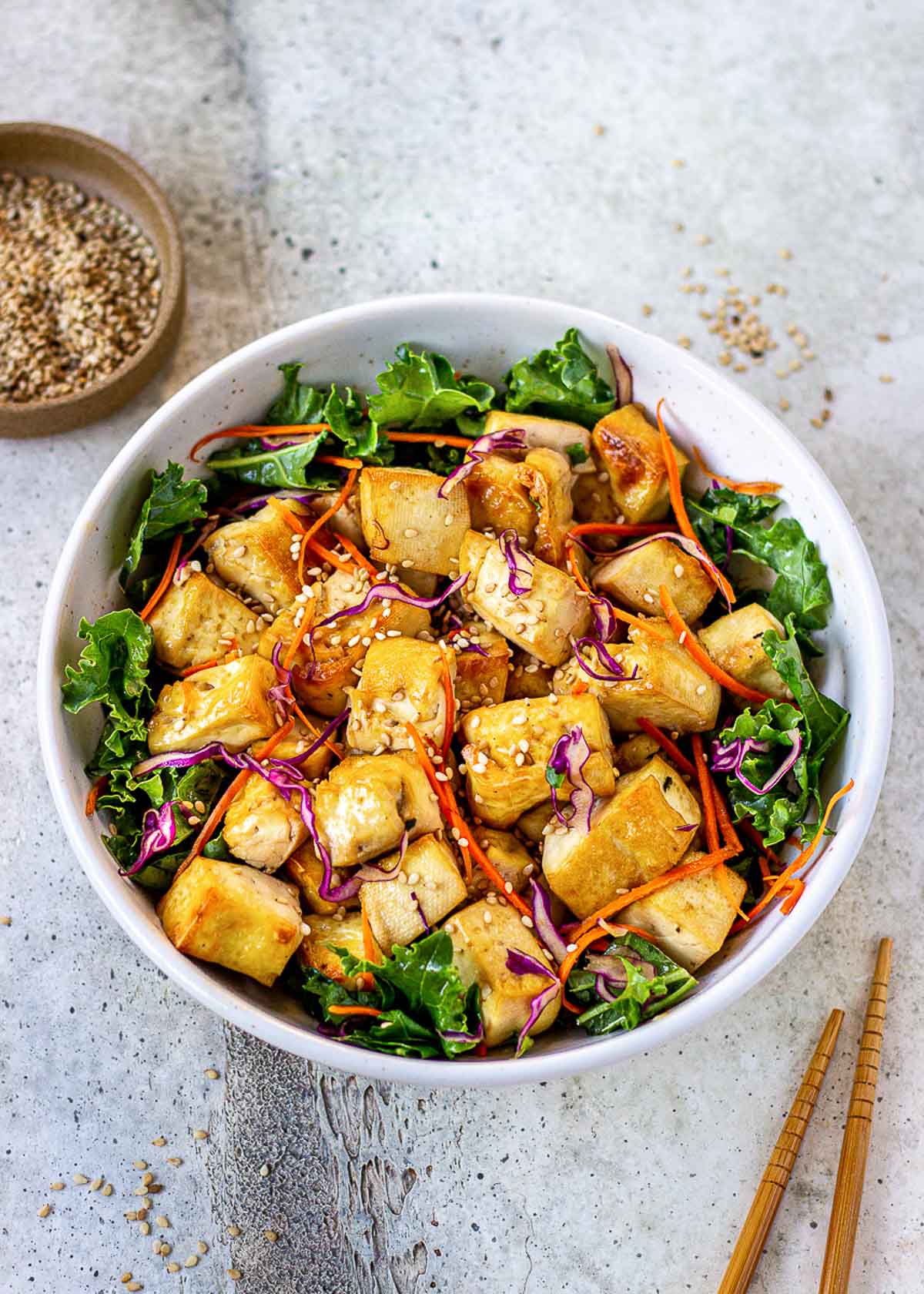 Bowl of healthy tofu surrounded by green leaves, red cabbage, carrots and a bowl of sesame seeds.