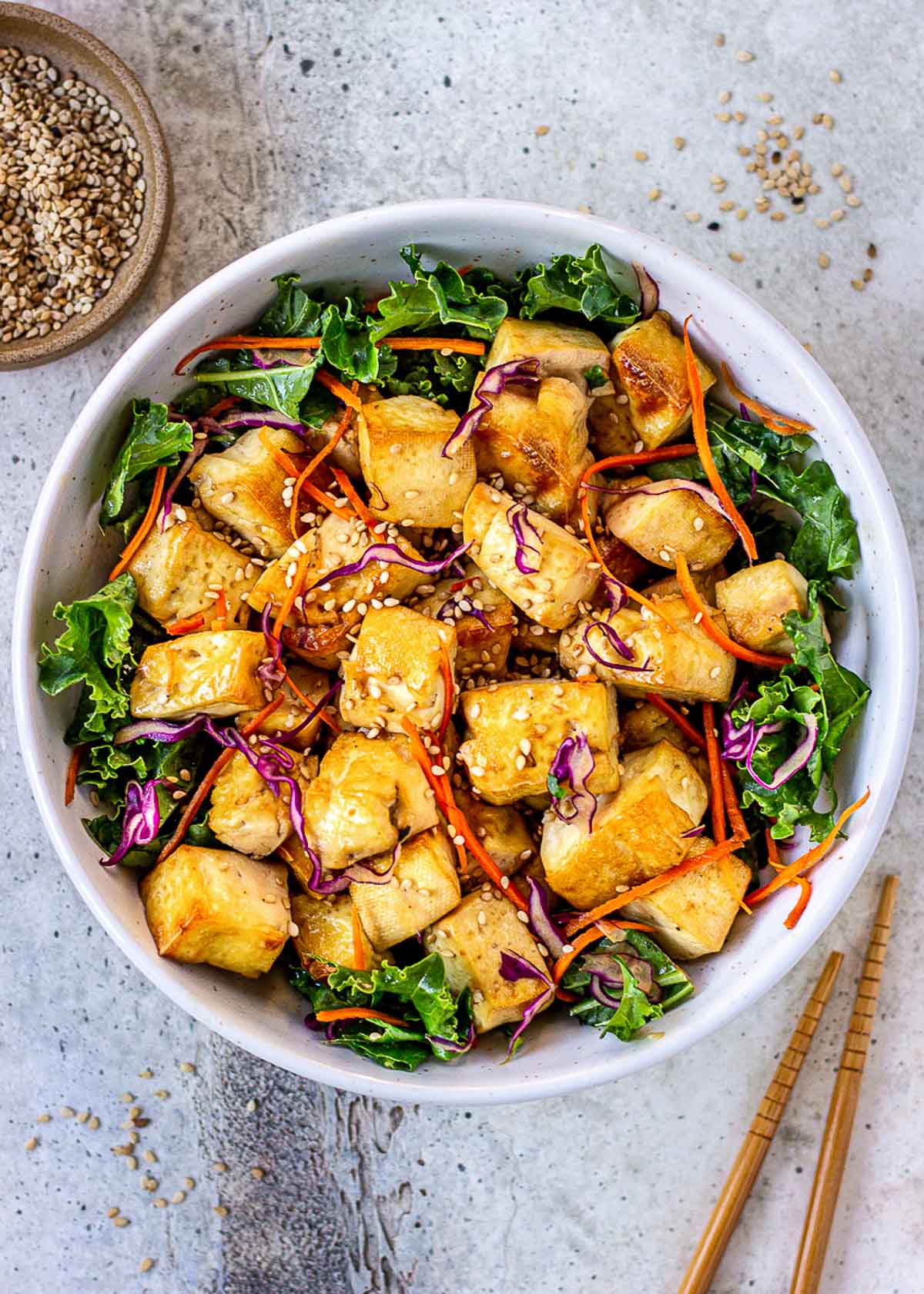 Bowl of broiled healthy tofu with kale, cabbage, carrot and sesame seeds. Chopsticks and bowl of sesame seeds nearby.