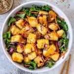 Bowl of crispy tofu surrounded by green leaves, red cabbage and carrots. Topped with sesame seeds.