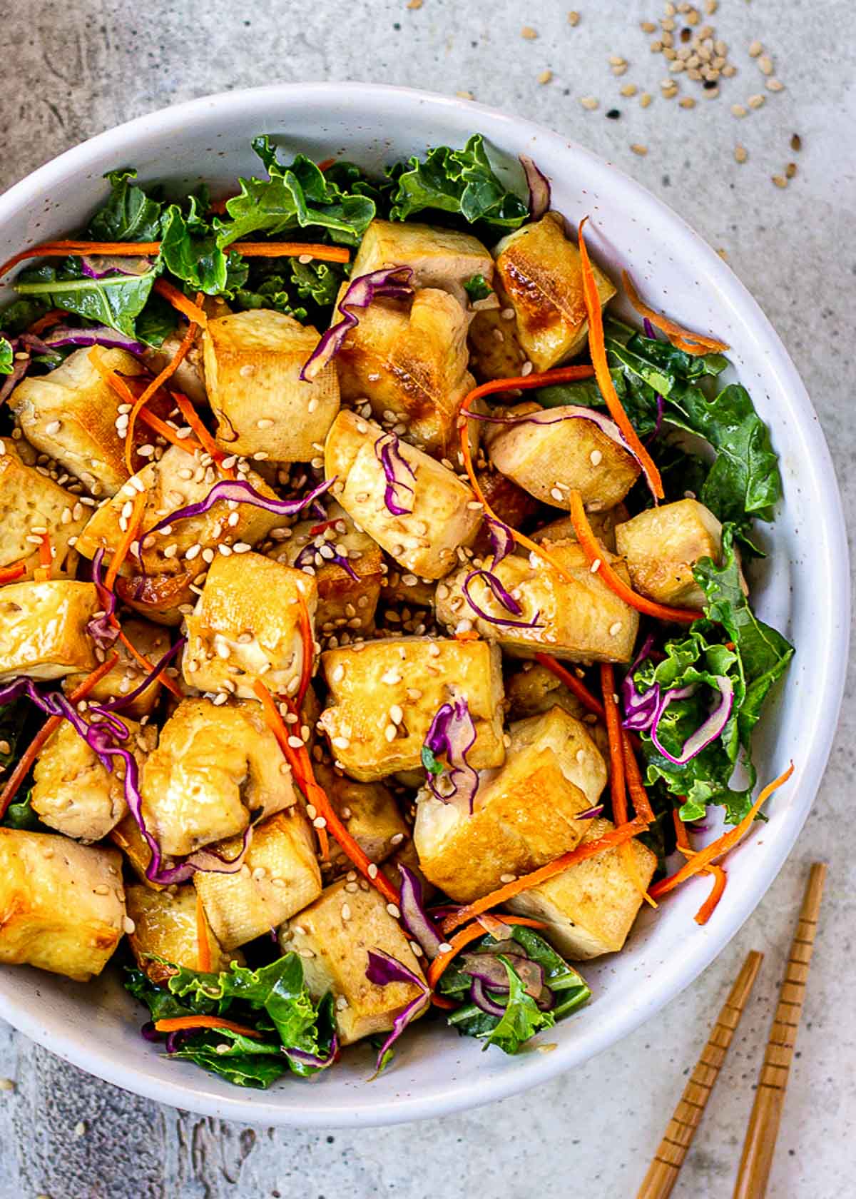 Bowl of crispy tofu surrounded by green leaves, red cabbage, carrots and chopsticks.
