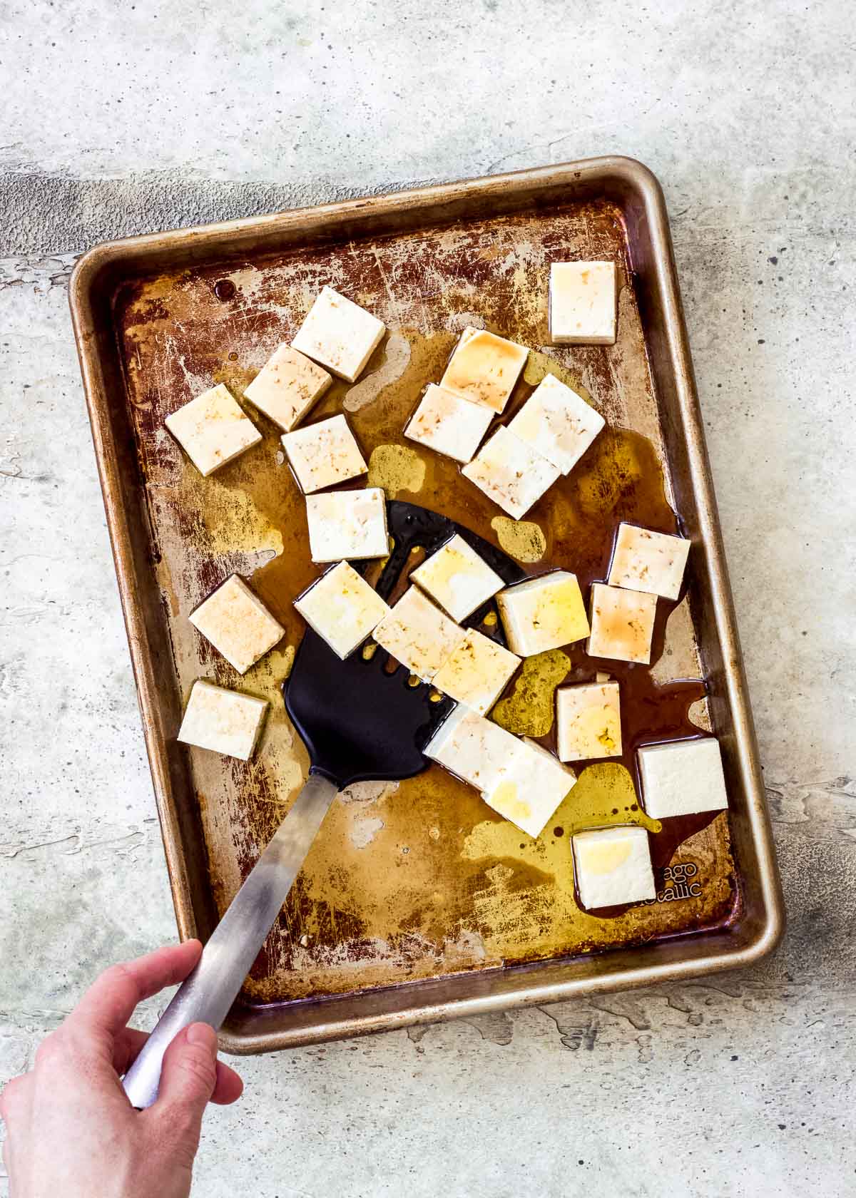 Tray of uncooked cubed tofu covered in oil and soy sauce, being flipped with spatula by female hand.