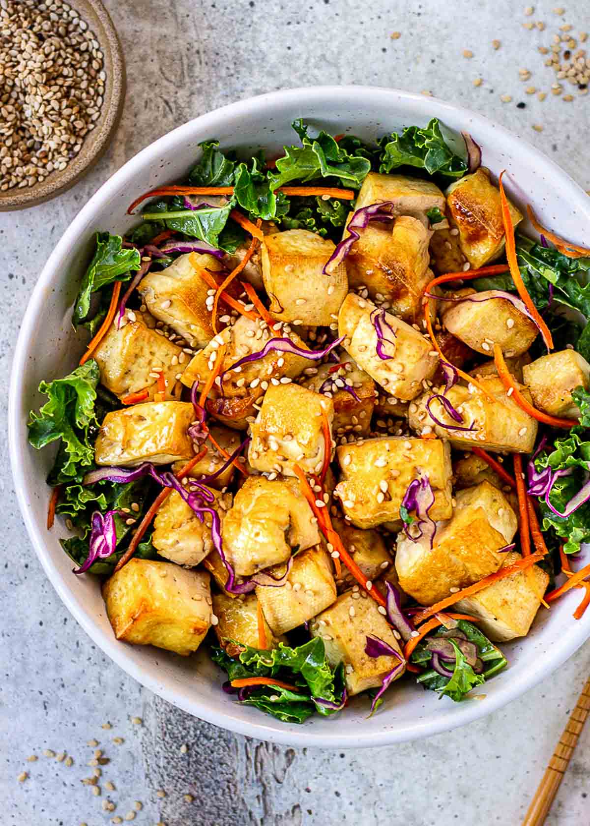 Bowl of crispy tofu surrounded by green leaves, red cabbage and carrots. Topped with sesame seeds.