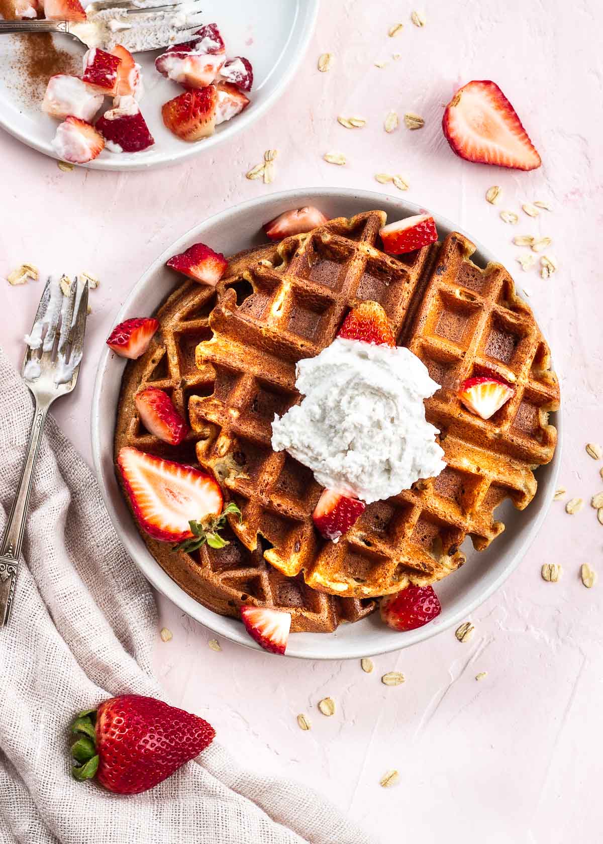 Oat Banana Waffles on a plate decorated with strawberries and coconut whipped cream, with a fork off to one side.
