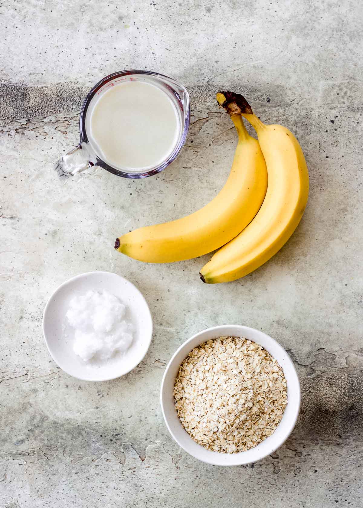 Oats, banana, milk and coconut oil on a grey surface.