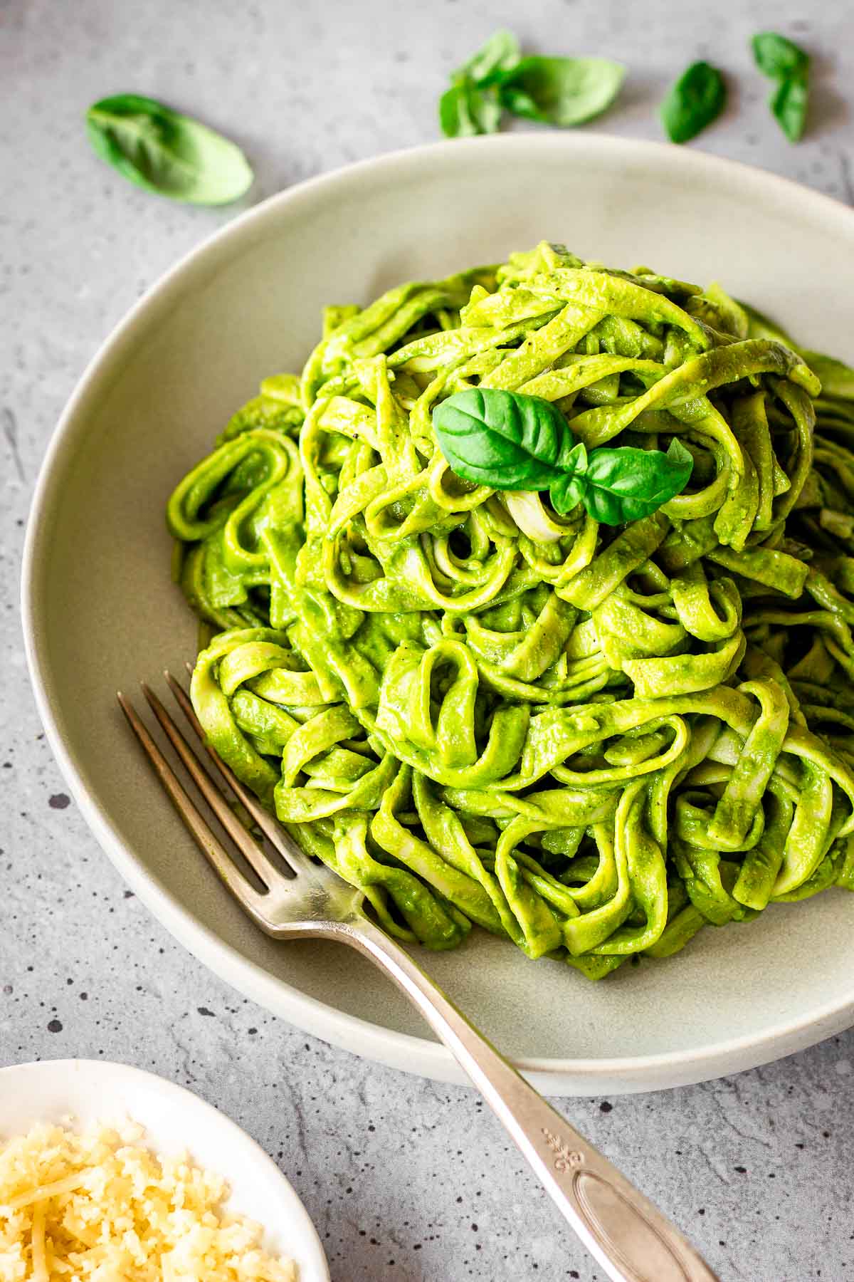Grey plate of creamy spinach pasta sauce on fettuccine. Dish is decorated with basil leaves and there is a fork nestled into the plate. A dish of vegan cheese is off to one side.