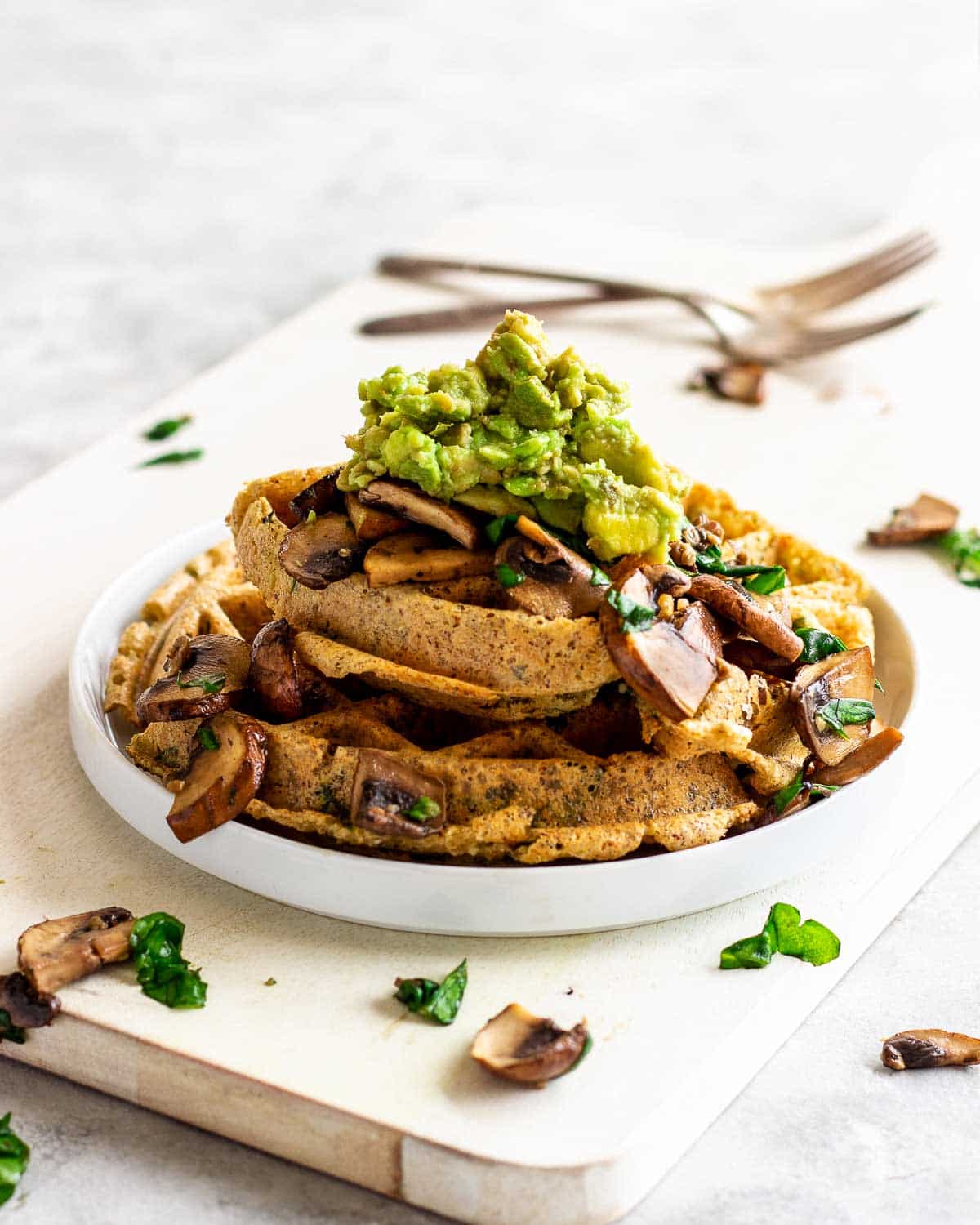 Vegan spinach waffles with mushrooms and avocado, on a white chopping board with a fork nearby.