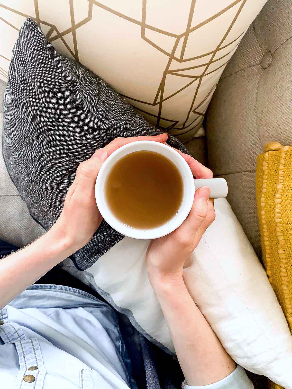 Overhead shot of a woman's hands holding a mug of tea on a sofa, surrounded by cushions.