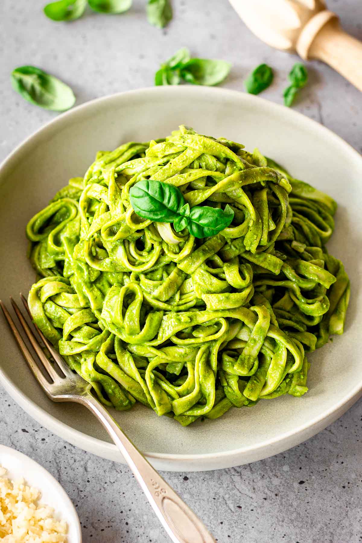 Grey plate of vegan creamy spinach pasta sauce on fettuccine. Dish is decorated with basil leaves and there is a fork nestled into the plate. A dish of vegan cheese is off to one side.