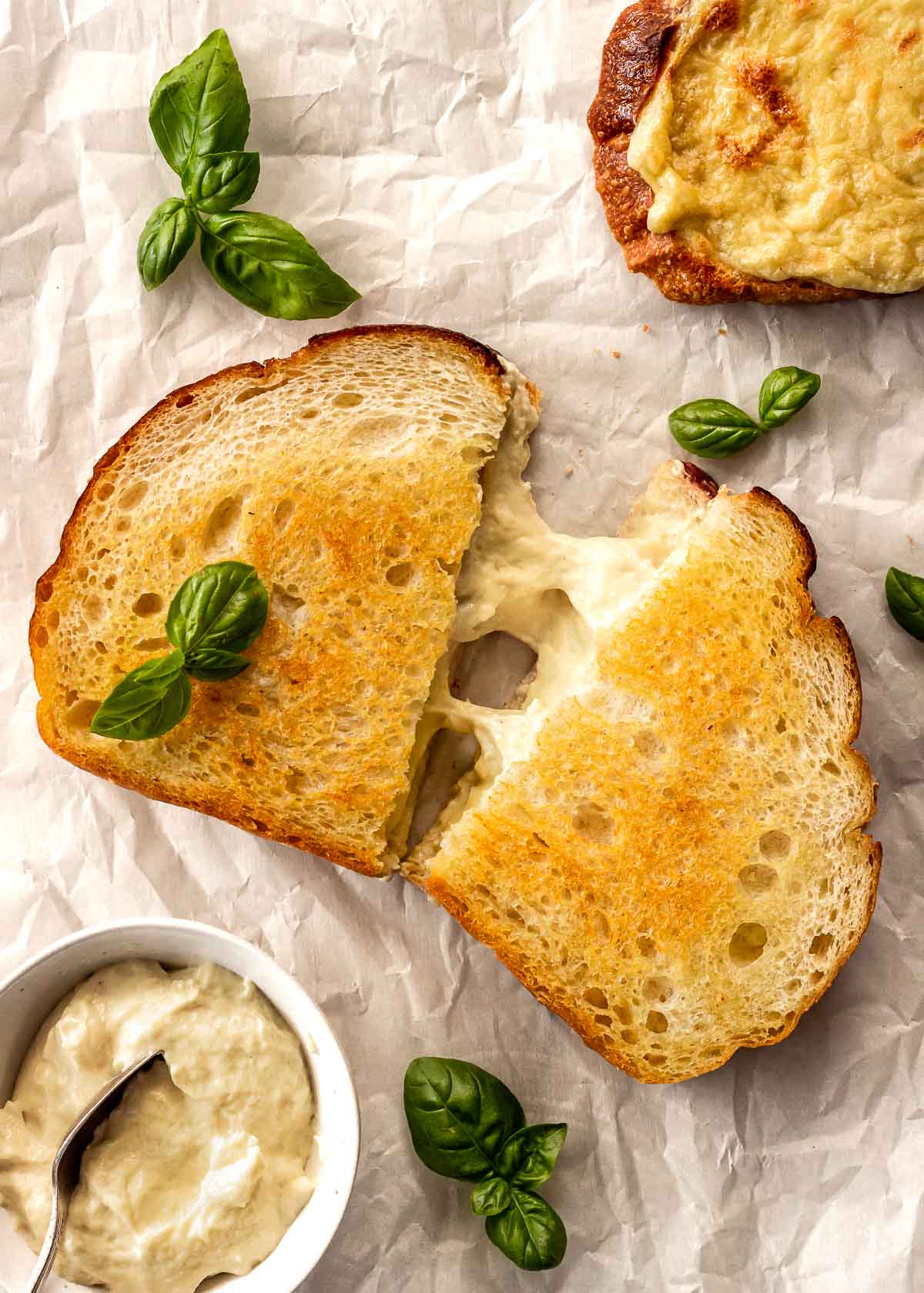 Vegan mozzarella shown in a grilled cheese sandwich with basil leaves scattered around.