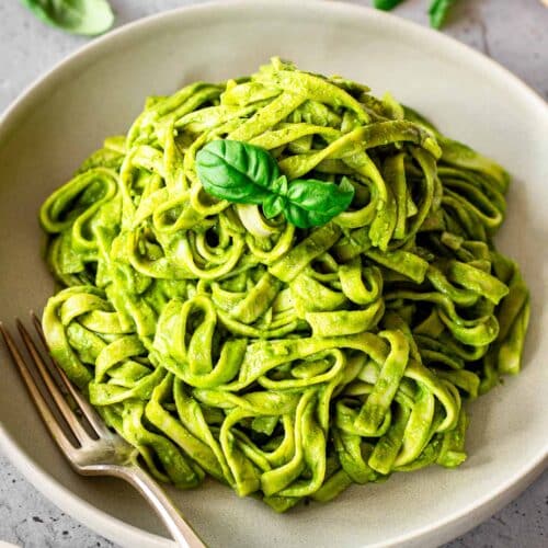 Grey plate of vegan creamy spinach pasta sauce on fettuccine. Dish is decorated with basil leaves and there is a fork nestled into the plate.