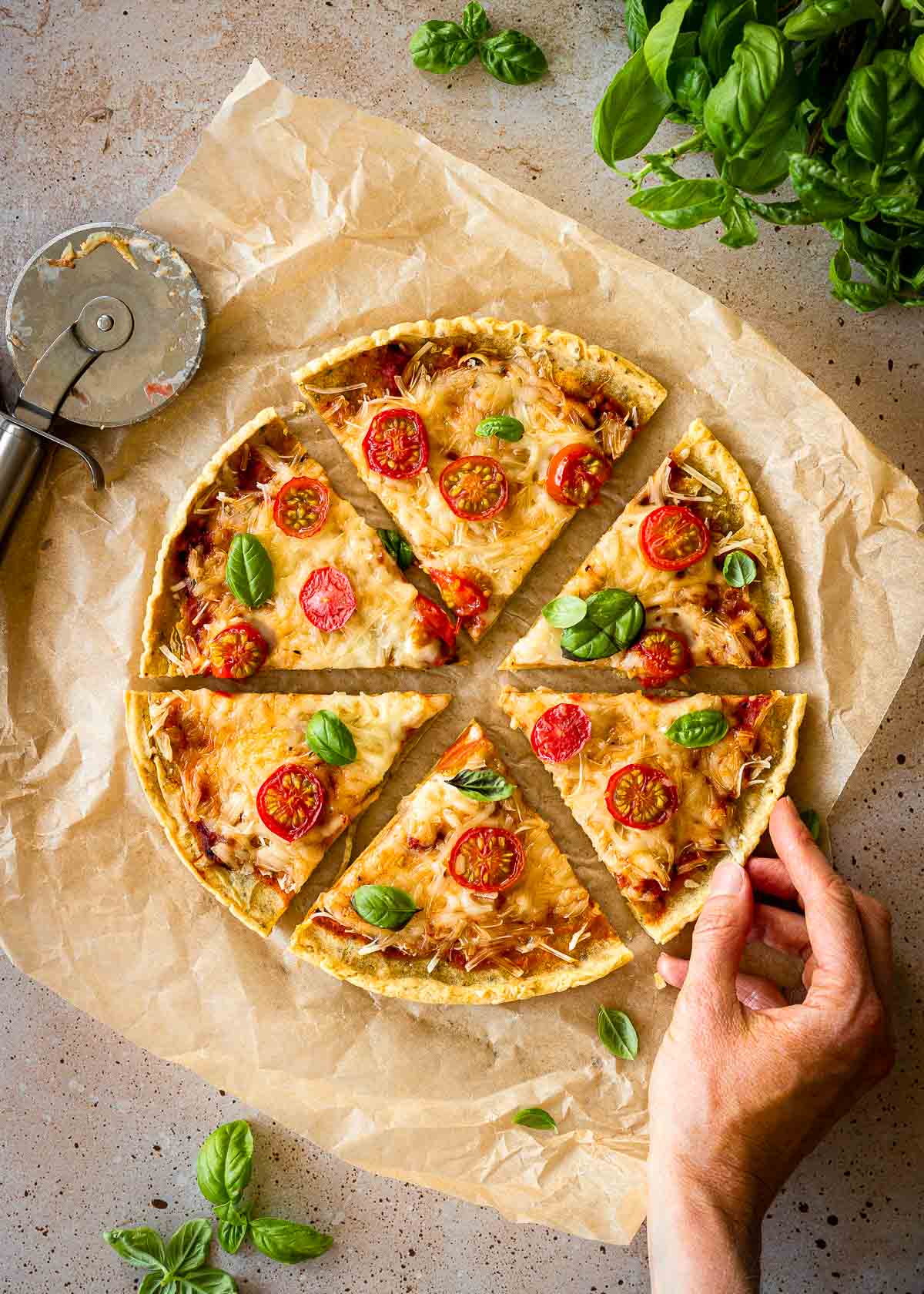 A woman's hand takes a slice of chickpea pizza on parchment paper. Basil leaves and a pizza wheel sit nearby.