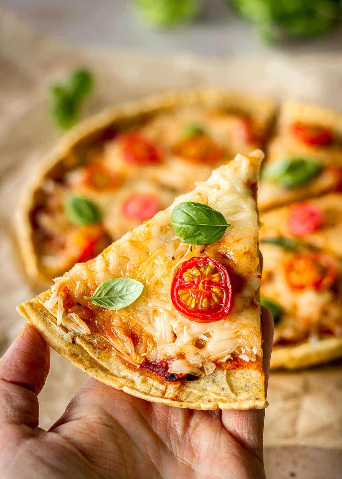 A woman's hand holds a slice of chickpea flour pizza, topped with cheese, cherry tomatoes and basil leaves. The rest of the pizza is in the background.