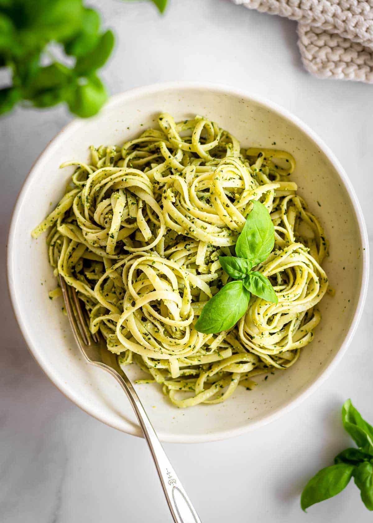 White bowl of pasta al pesto with basil leaves and on top and a fork on the side. More basil leaves and a white cloth are off to the side.