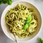 White bowl of pasta al pesto with basil leaves and on top and a fork on the side.