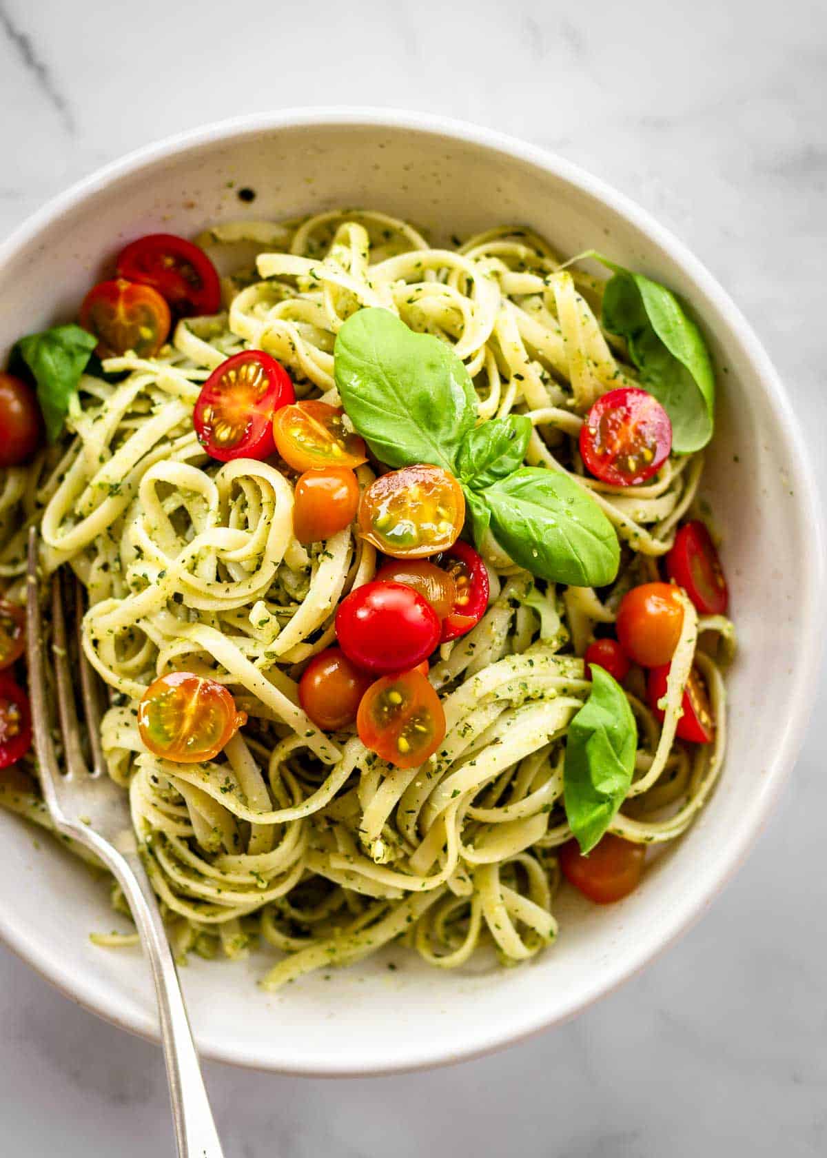 White bowl of pasta al pesto with basil leaves and cherry tomatoes on top.